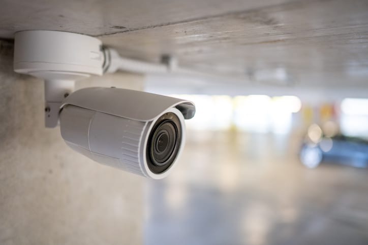 Surveillance camera mounted under a concrete ceiling in a dim parking garage, monitoring the area.